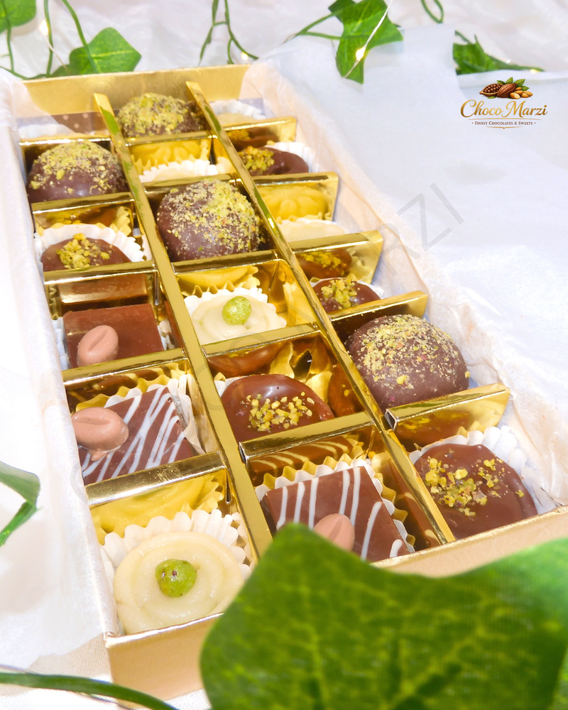 A gold-lidded box of assorted chocolates arranged in a grid, photographed on white tissue paper with green leaves around the edges. The chocolates include dark chocolate truffles with gold sprinkles, milk chocolate squares with white drizzle, and cream-colored chocolates with green centers.