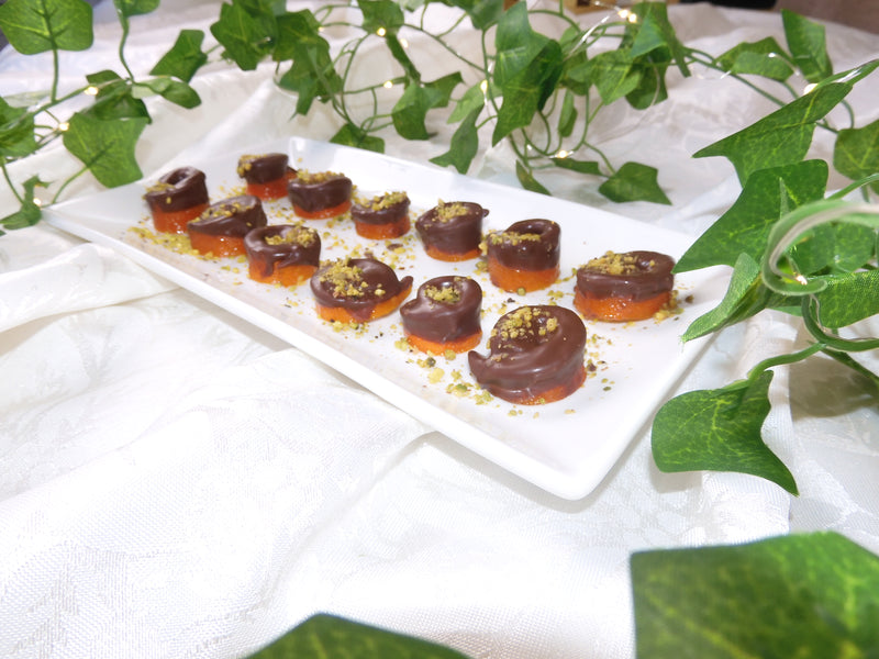 A white rectangular plate holding several small rolled appetizers or candies, each with a dark brown top and a translucent orange-red base, garnished with crushed green nuts. The plate is styled on a white fabric surface with green ivy leaves around it.